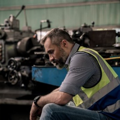 A bearded man in a short and high vis vest sits with his head bowed in a factory.